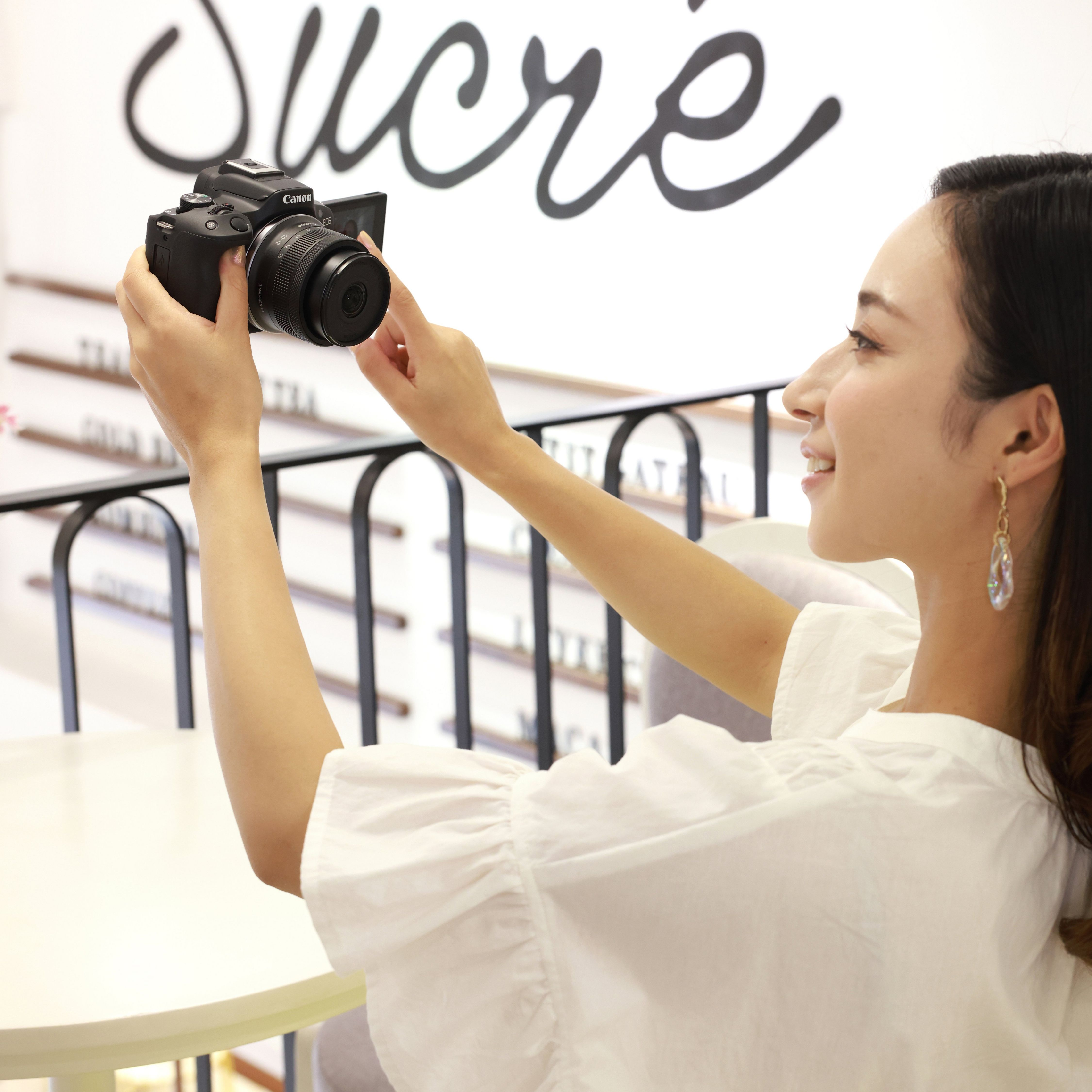 Une femme prend un selfie avec un appareil photo dans un café. Un panneau indique 'Sucre'.