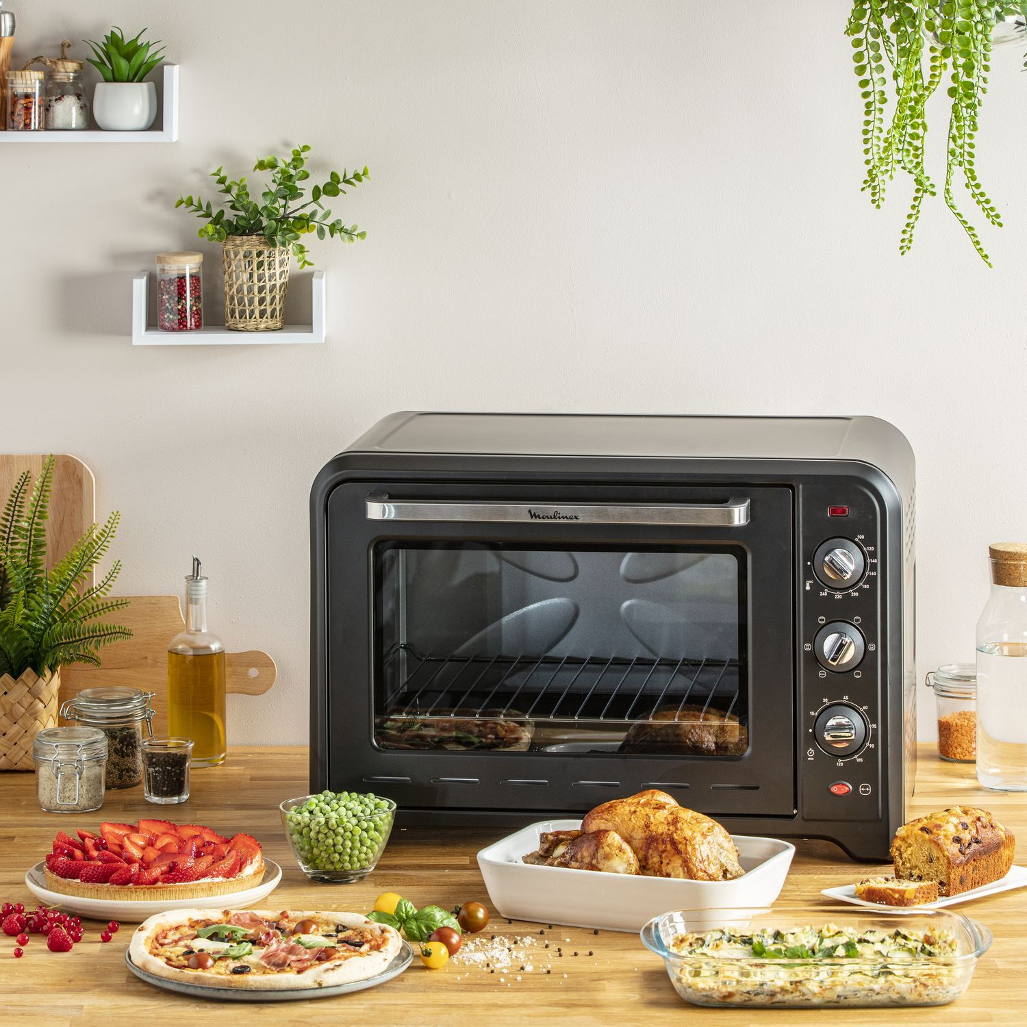 A black oven with food on the counter. Includes a whole chicken, pizza, and vegetables. Plants in the background.