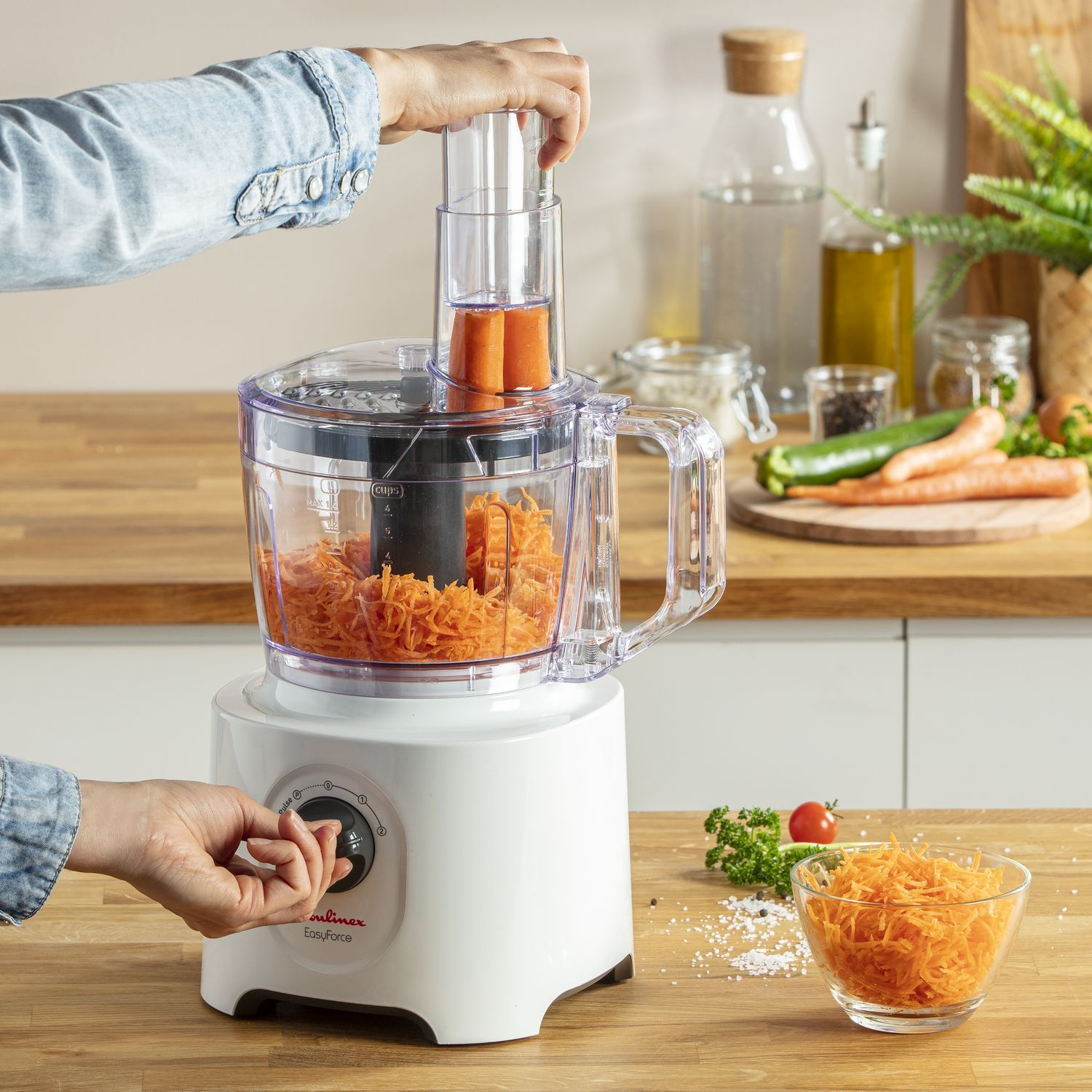 Personne utilisant un robot culinaire pour râper des carottes. Un bol en verre avec des carottes râpées est sur le comptoir.
