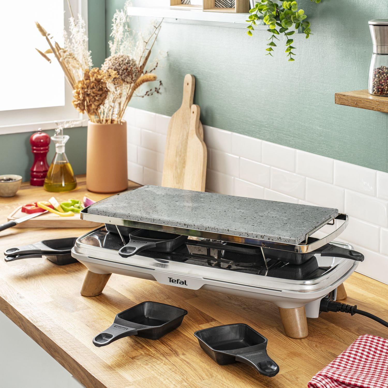 Tefal raclette grill met pannetjes op een houten tafel. Voedsel en serveerartikelen zijn aanwezig. Groene muurachtergrond.