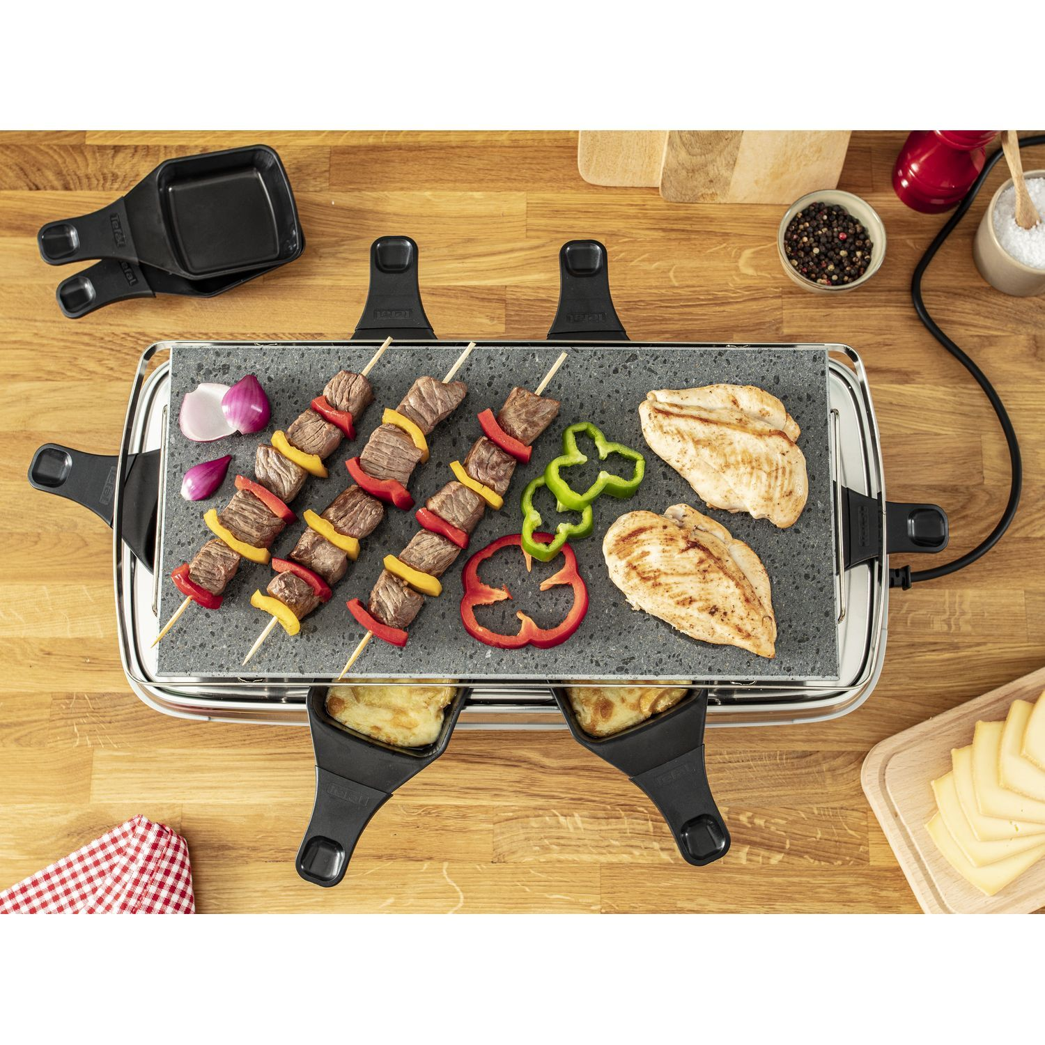 Een grill met vlees en groenten op een houten tafel, inclusief gereedschap.