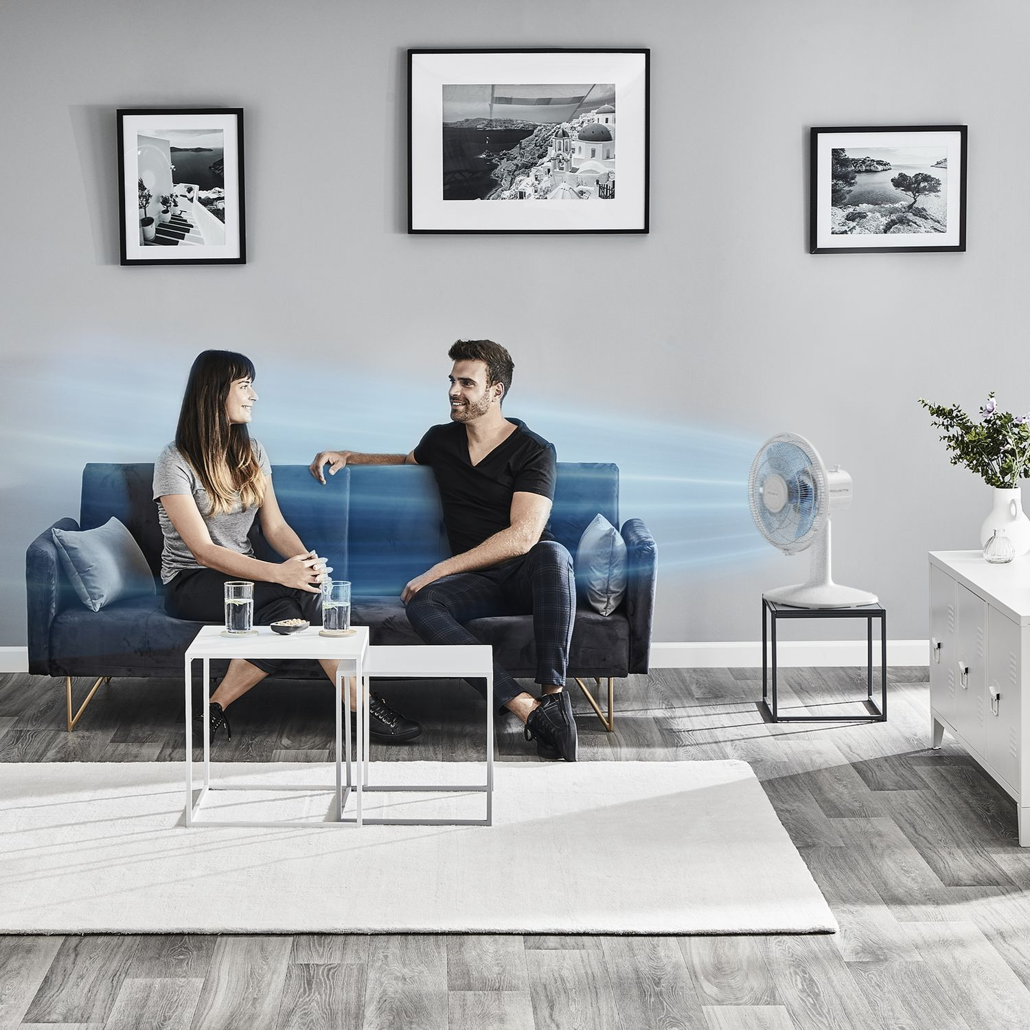 A couple sitting on a couch with a fan blowing air.