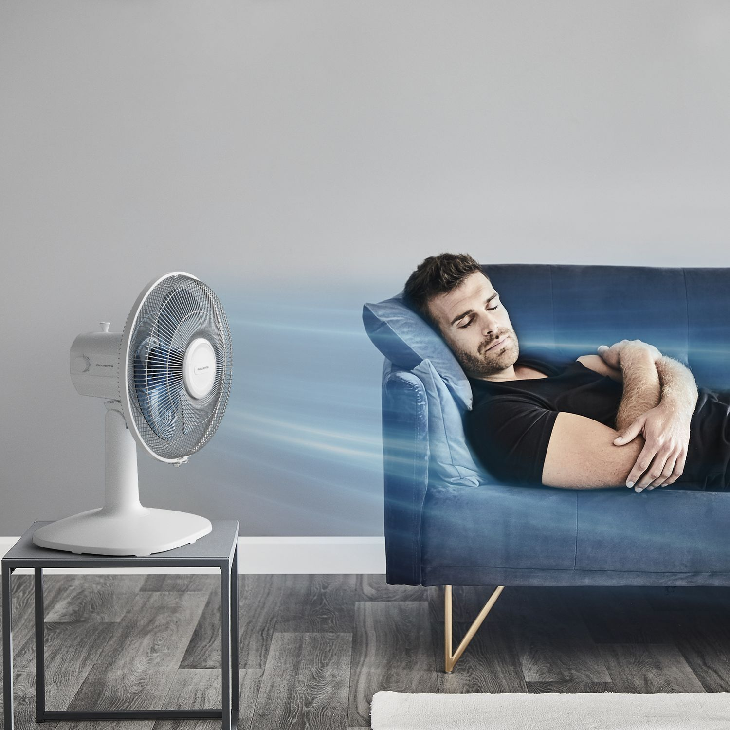 A man sleeping on a blue couch, a white fan is to the left, blowing air towards him.