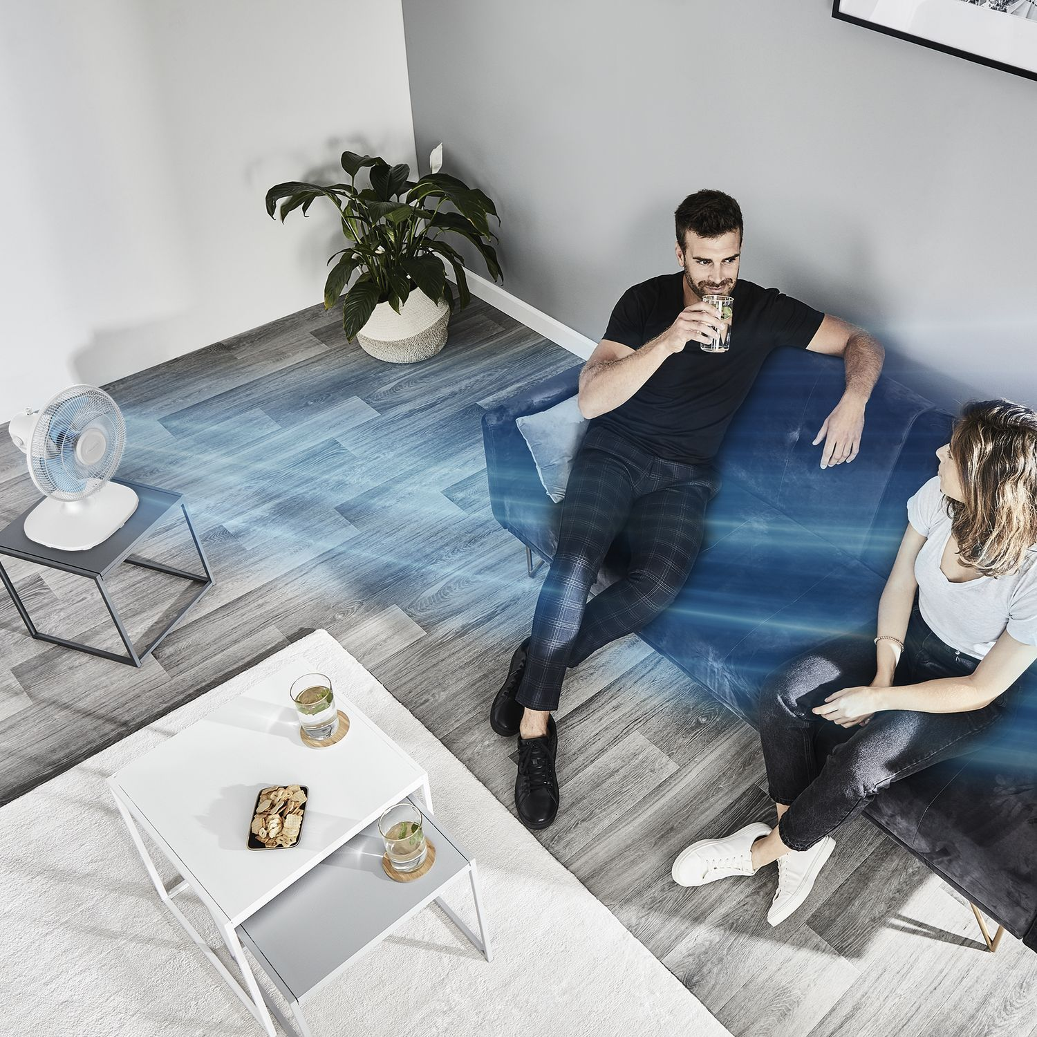 Two people sitting on a couch, with a fan blowing air. Two tables are in front.