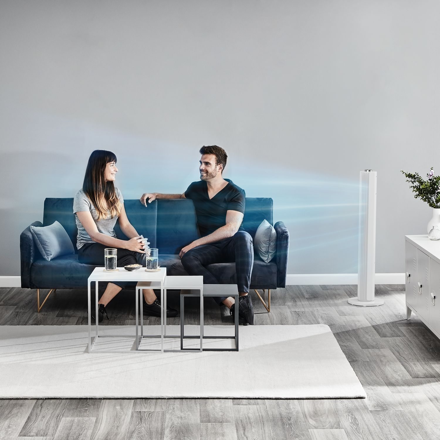 Een stel zit op een blauwe bank en kijkt naar een witte ventilator. Twee kleine tafels en een tapijt zijn zichtbaar.
