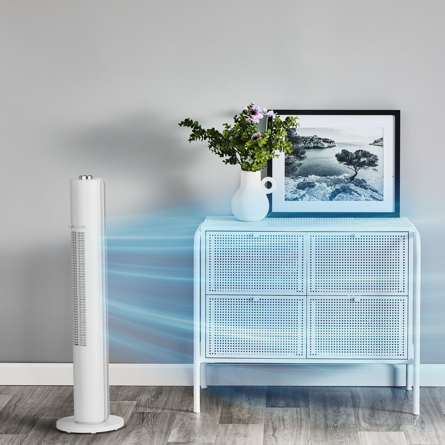 Een witte ventilator die lucht blaast, naast een witte kast met een foto en een vaas met bloemen.