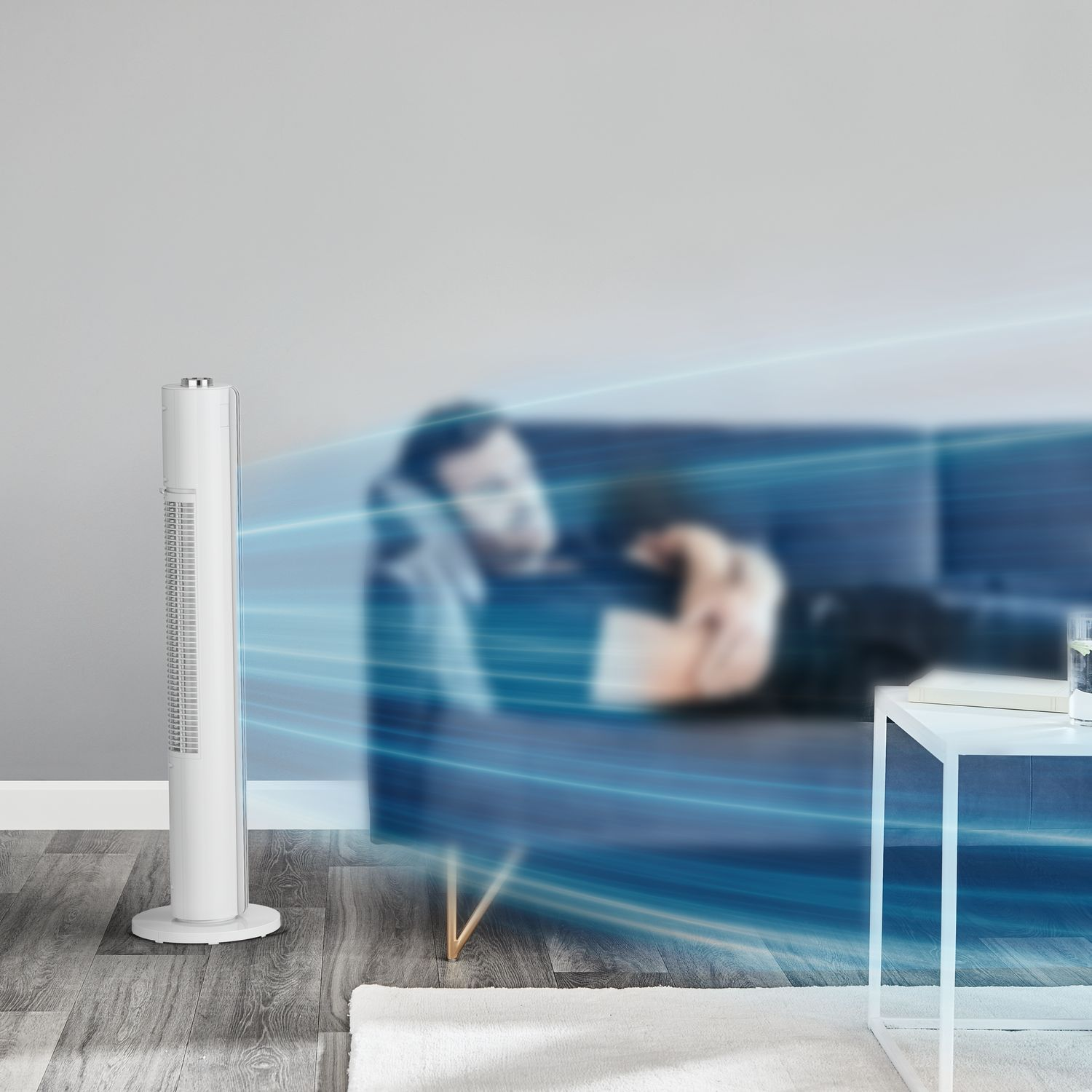 Een witte torenventilator in een kamer met een bank, persoon wazig op de achtergrond.