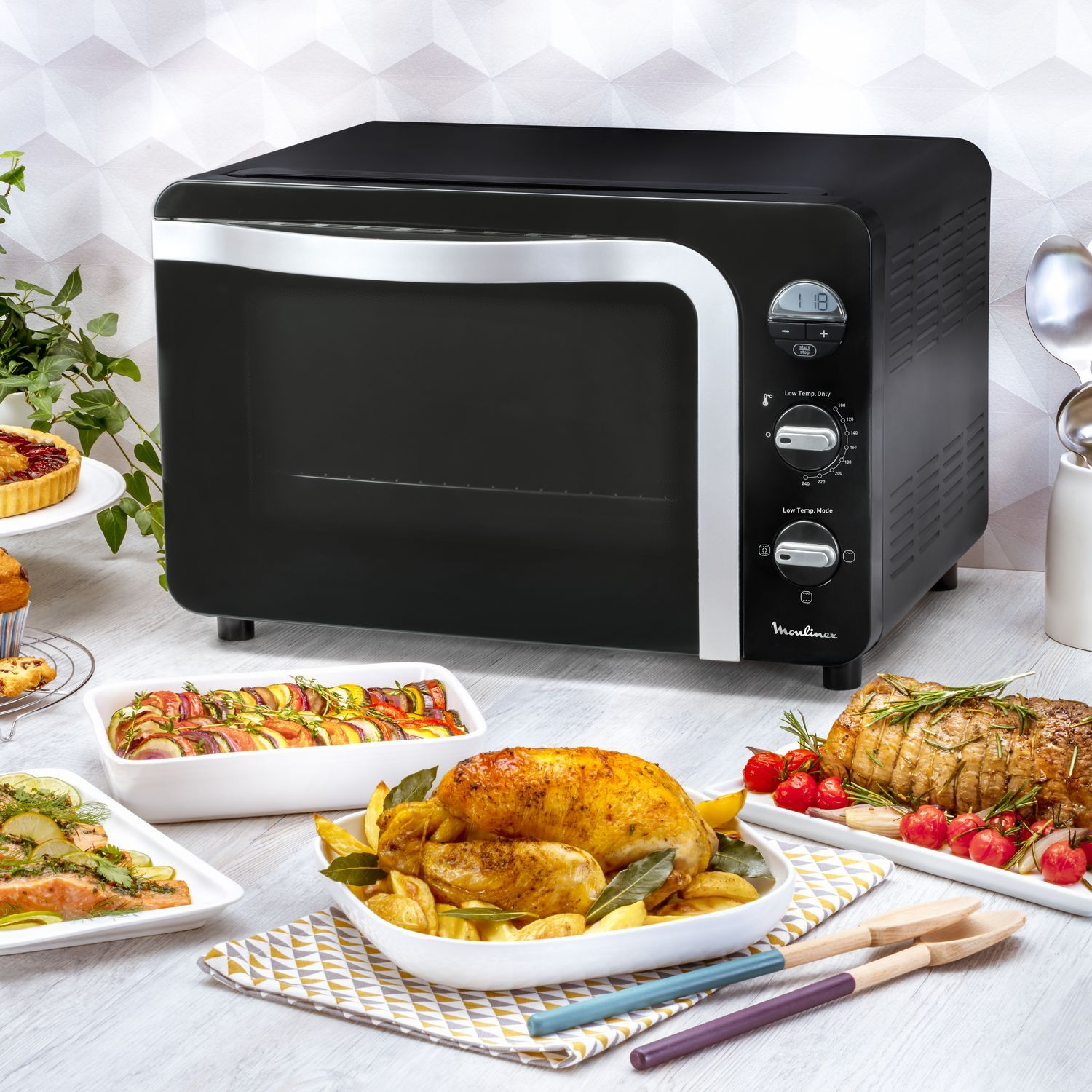 Zwarte Moulinex-oven met eten op tafel: gebraden kip, ratatouille, taarten en zalm.