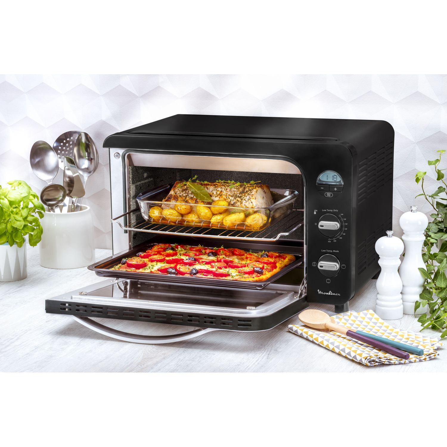 Zwarte oven met eten erin, deur open en keukengerei aan de zijkant.