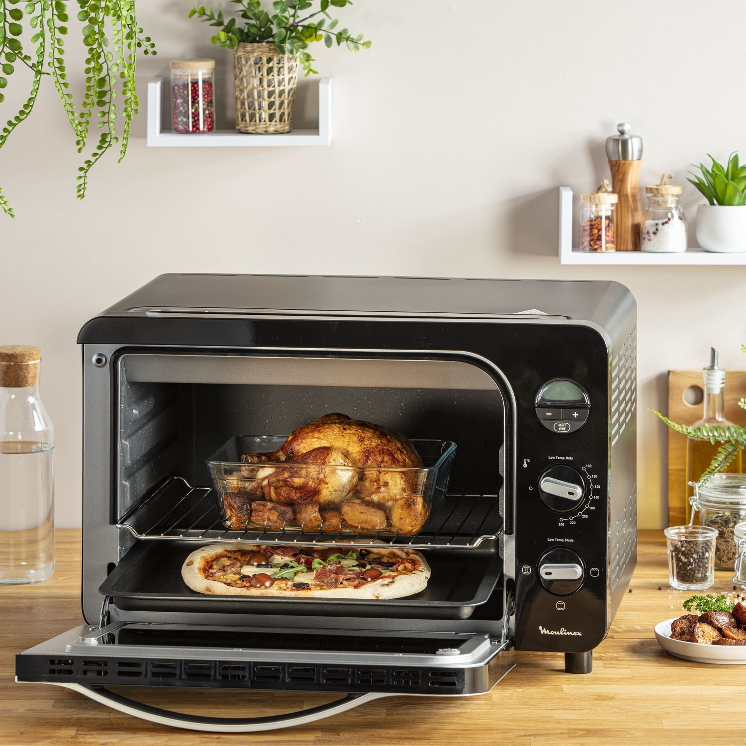 Een zwarte Moulinex oven met een gebraden kip en een pizza. Plankjes, potten en een houten tafel op de achtergrond.