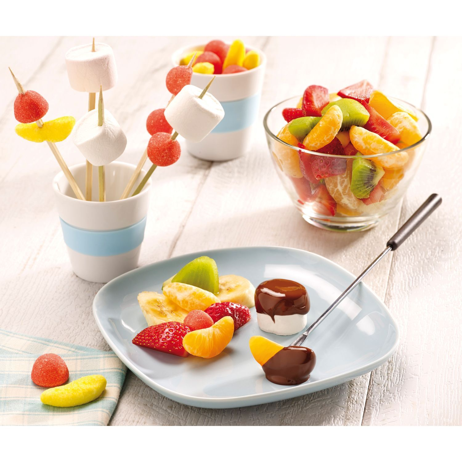 Ensemble fondue de fruits. Brochettes de fruits, guimauves et un bol de fruits. Assiette bleue et fond blanc.