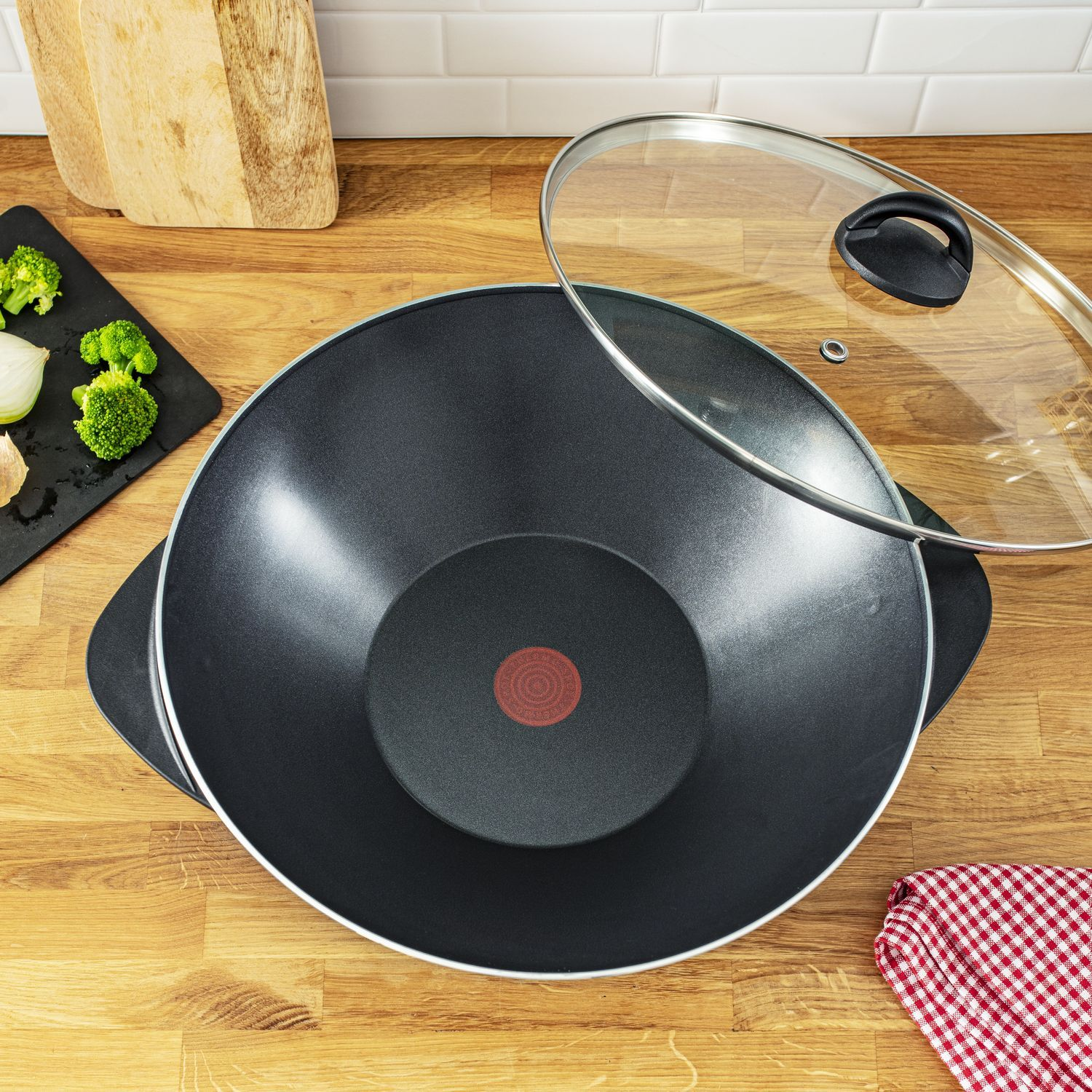 A black wok with a glass lid on a wooden surface, with ingredients and a cloth nearby.