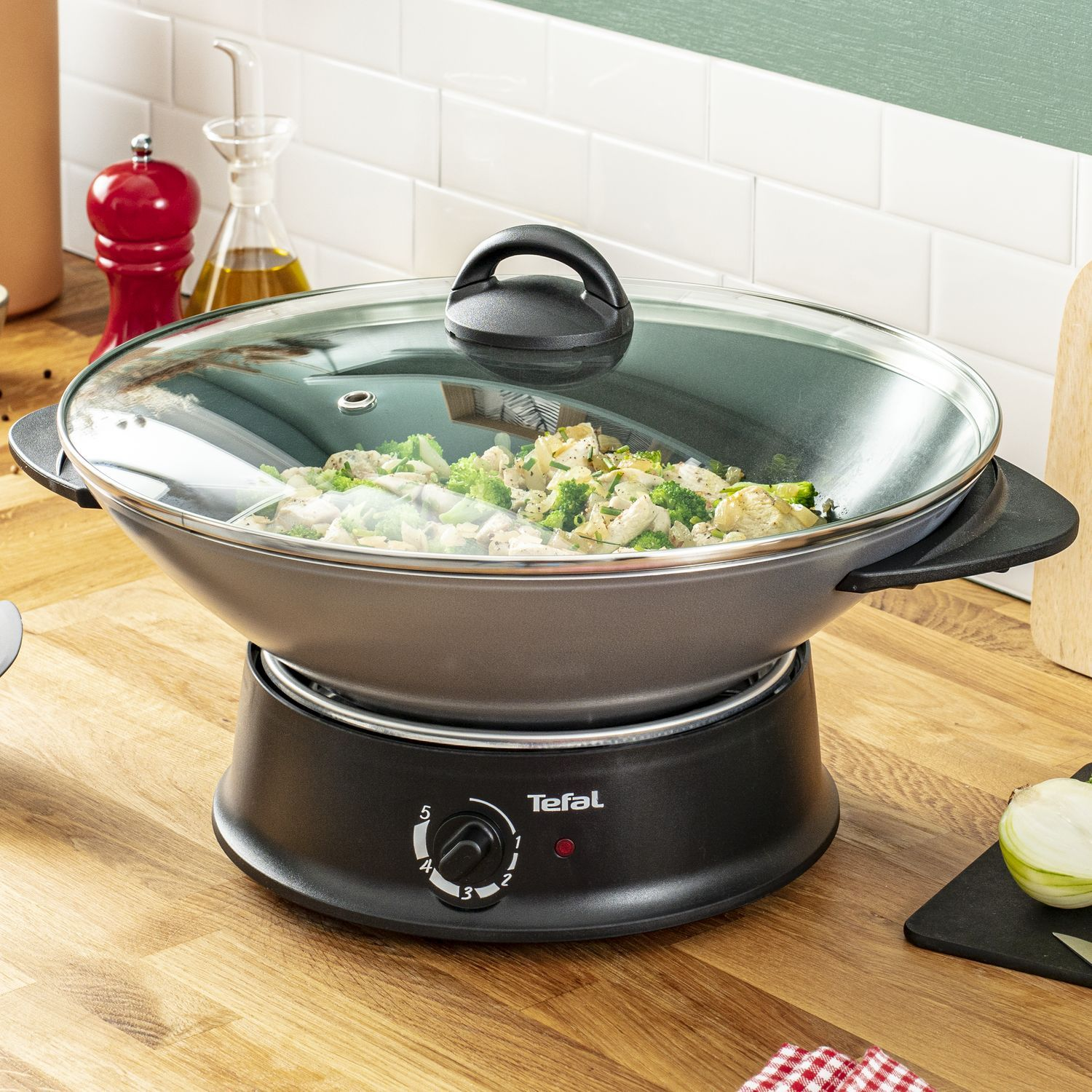 Black wok on electric stove. Glass lid. Vegetables visible. Wooden counter, white tiles.
