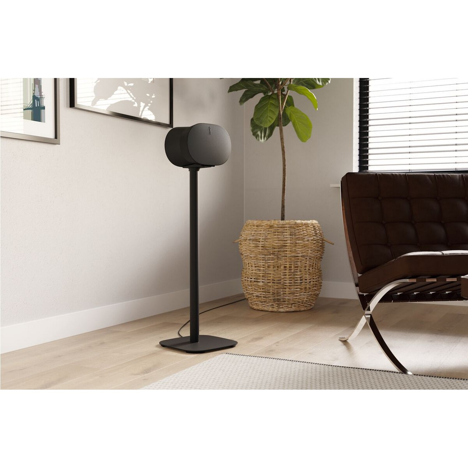 Een zwarte speaker op een standaard. Er is een plant in een mand, een stoel en foto's aan de muur.