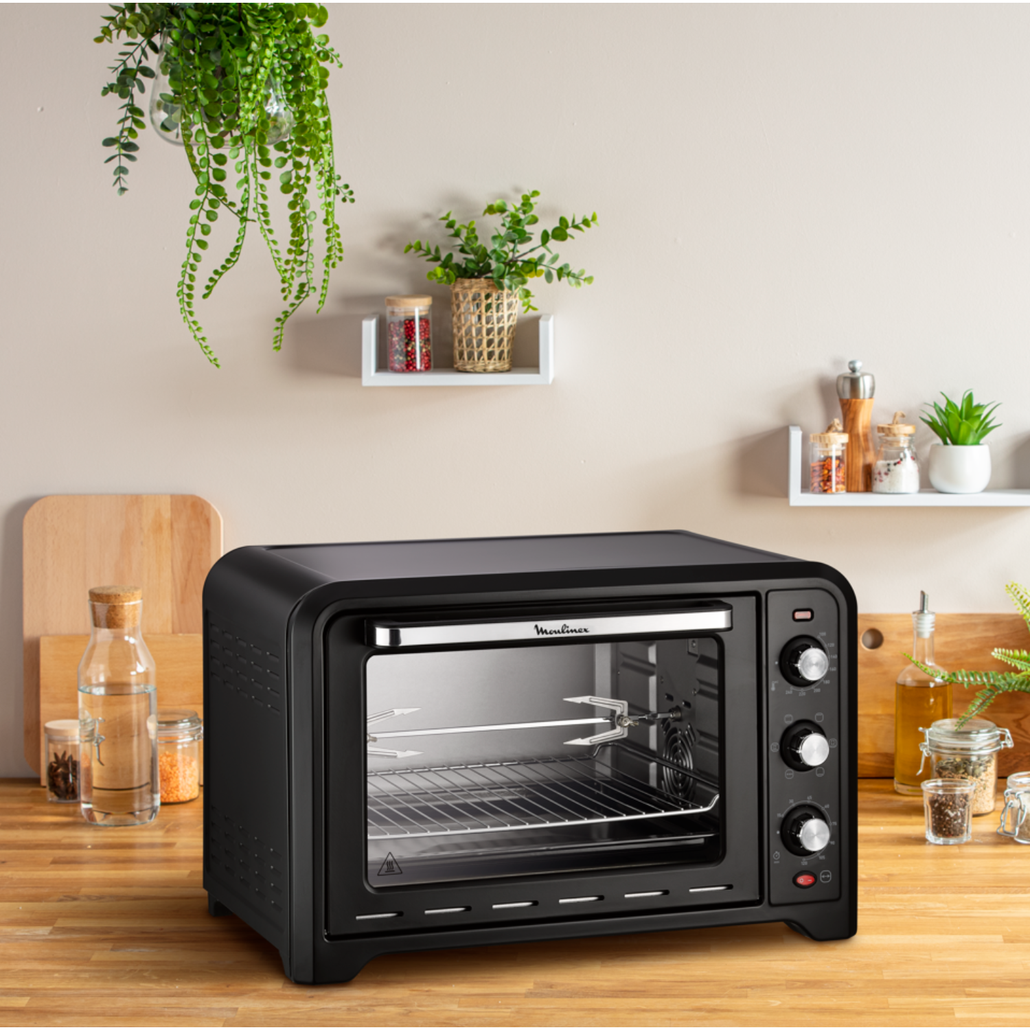 Black countertop oven with a glass door, on a wooden surface, plants overhead.