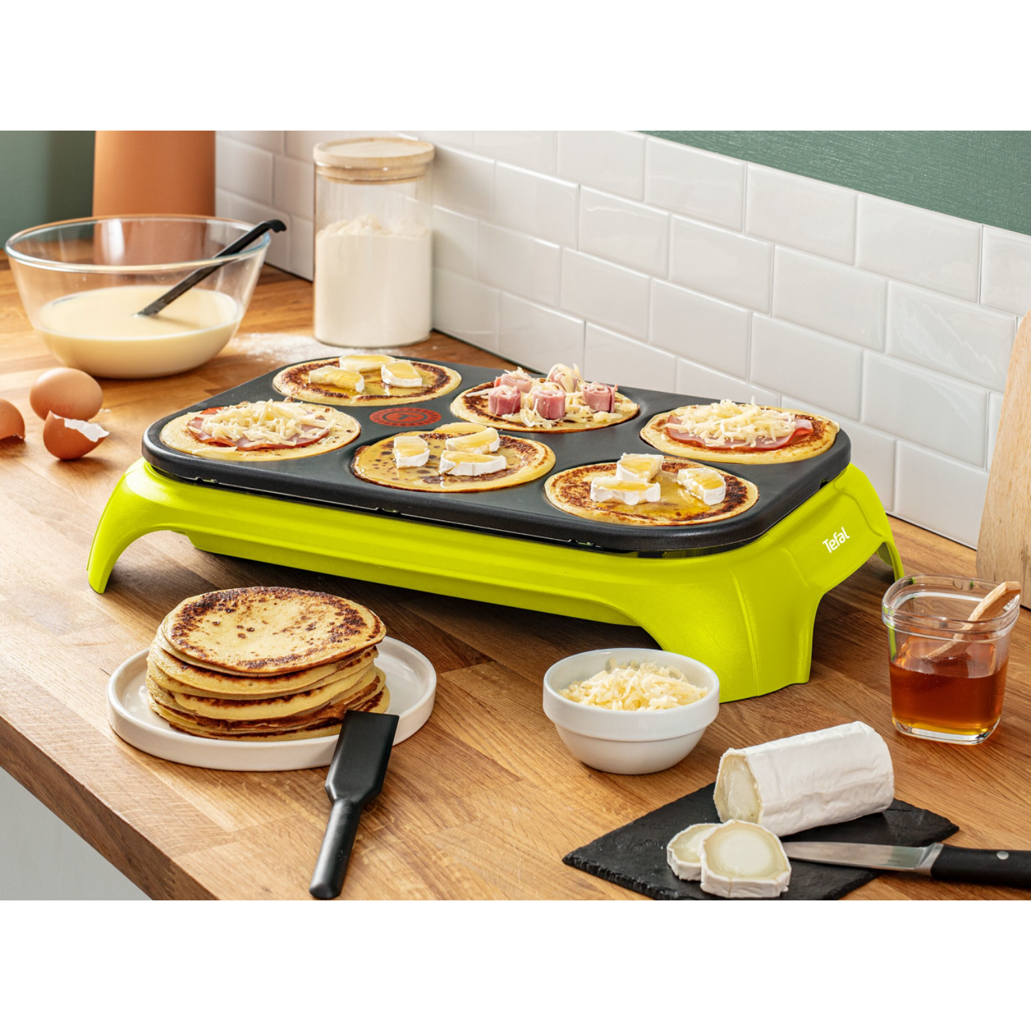Pancake maker with cooked pancakes, ingredients, and toppings arranged on a wooden surface.
