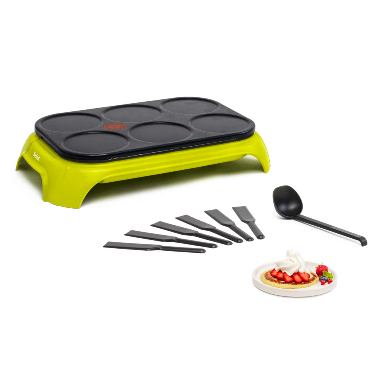A green pancake maker with a set of spatulas and a ladle. A plate with pancakes is in the background.