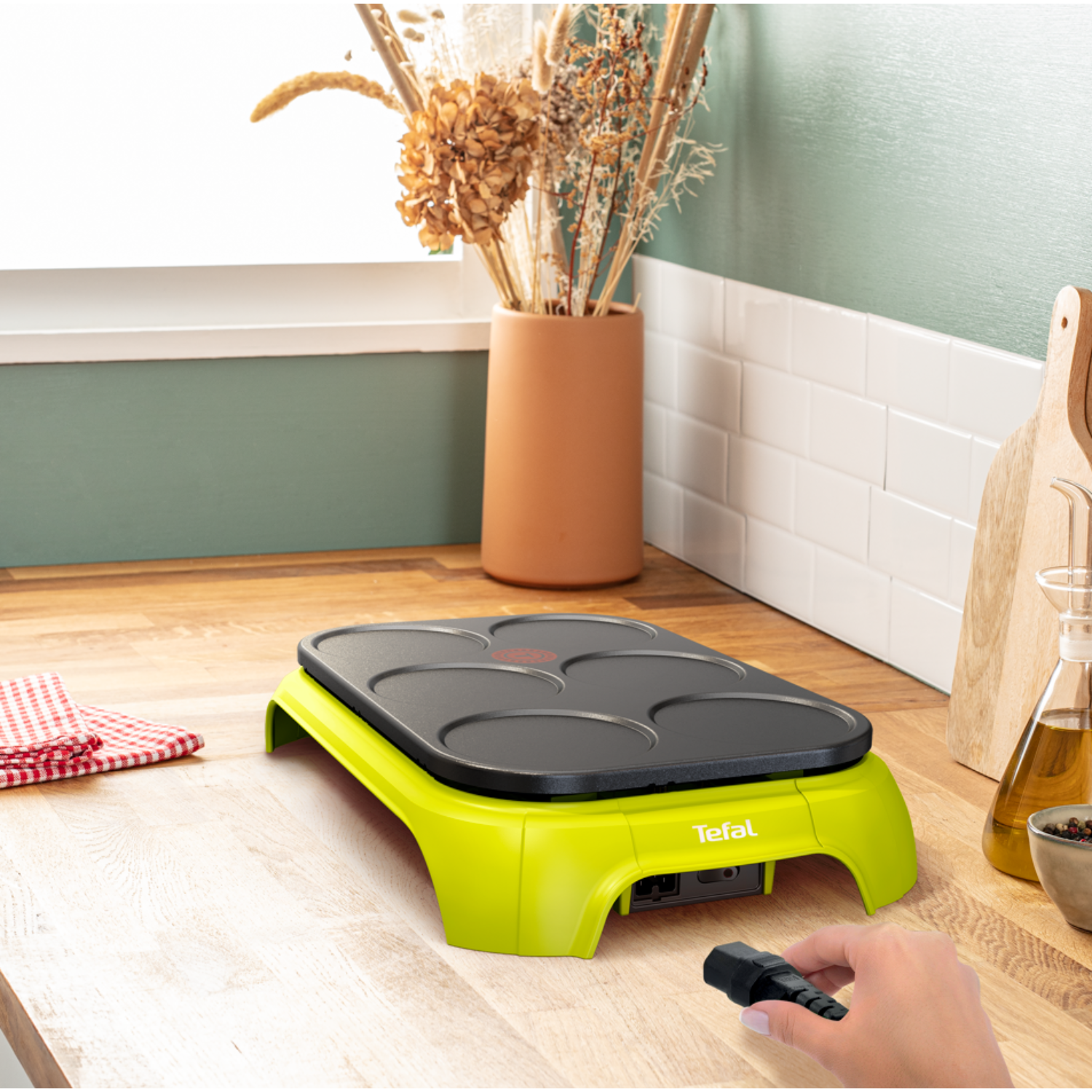A green crepe maker with a black handle and a hand.
