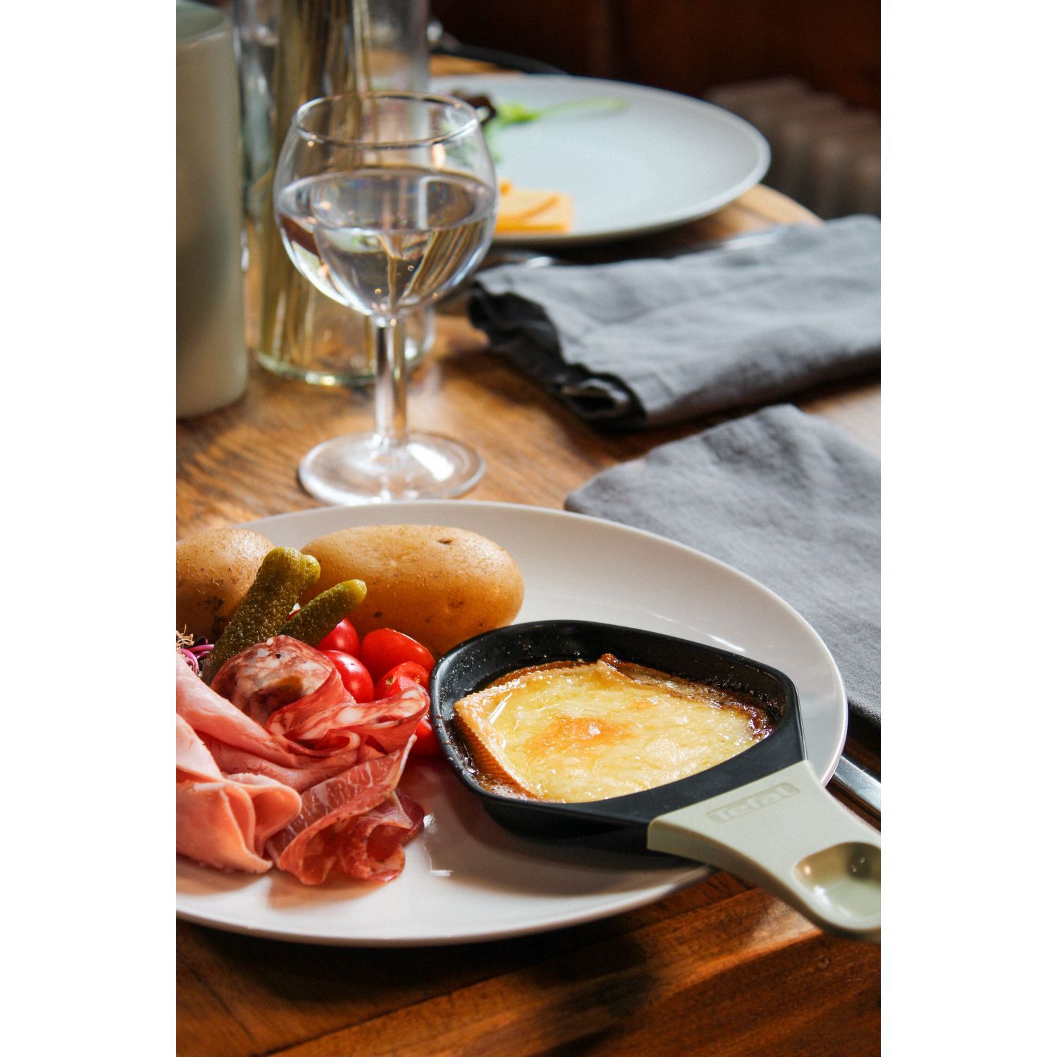 A plate with food: potatoes, pickles, ham, cheese. A glass of water. Background is wood.