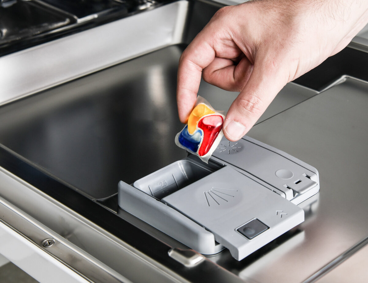 Ręka umieszcza wielokolorową kapsułkę do zmywarki w szarym dozowniku wewnątrz zmywarki ze stali nierdzewnej.