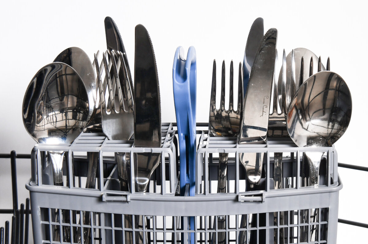 Sztućce w szarym koszu zmywarki. Widoczne widelce, noże i łyżki. Tło: białe.