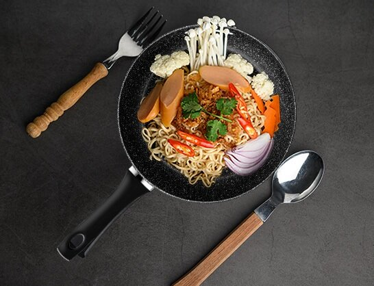 Patelnia z makaronem i dodatkami. Widelec i łyżka po bokach. Ciemna powierzchnia.