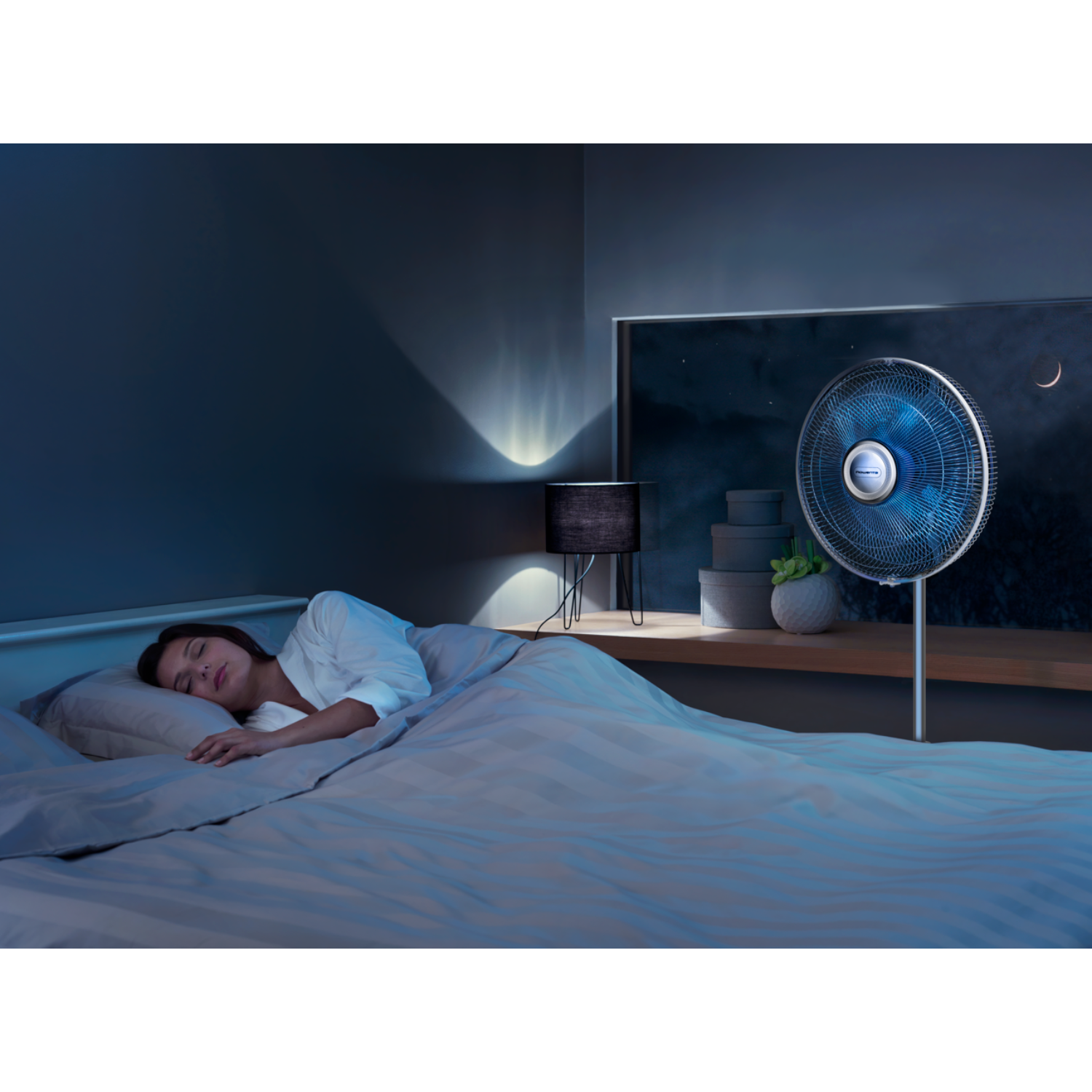 Woman sleeping in bed, with a fan on the right and a lamp.