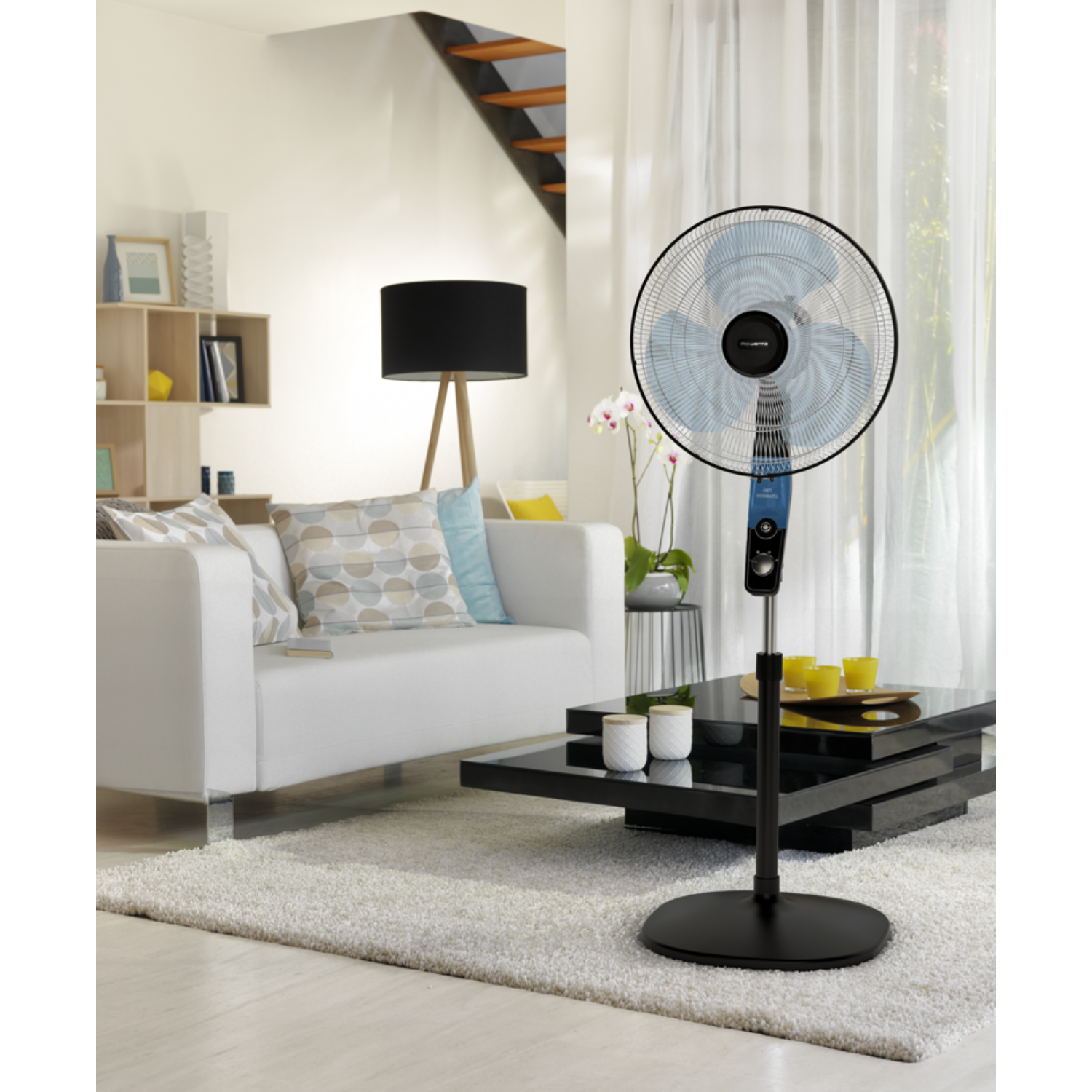 Un ventilateur sur pied dans un salon, canapé blanc, table basse noire et tapis blanc.