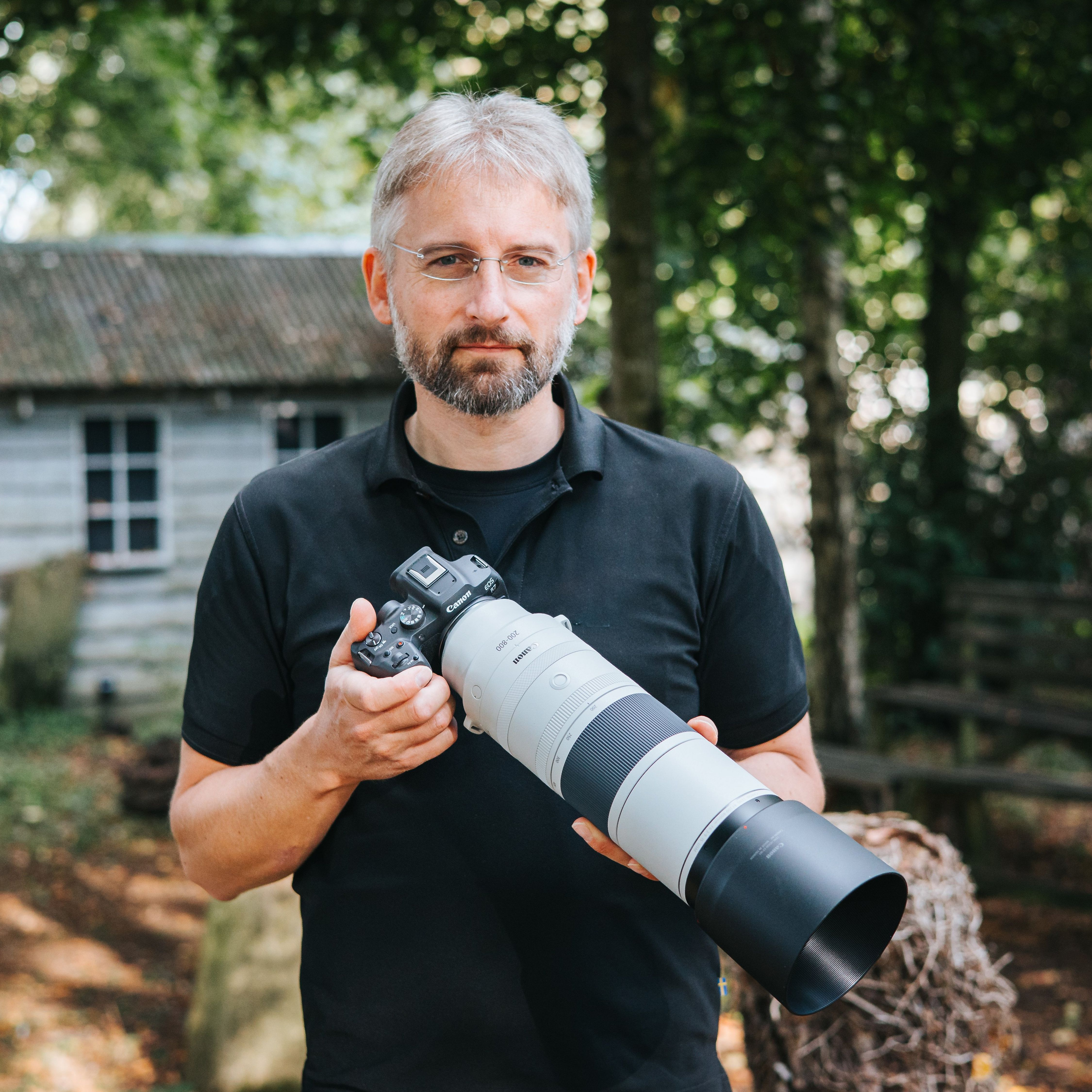 Un homme tenant un appareil photo avec un long objectif, devant une maison.