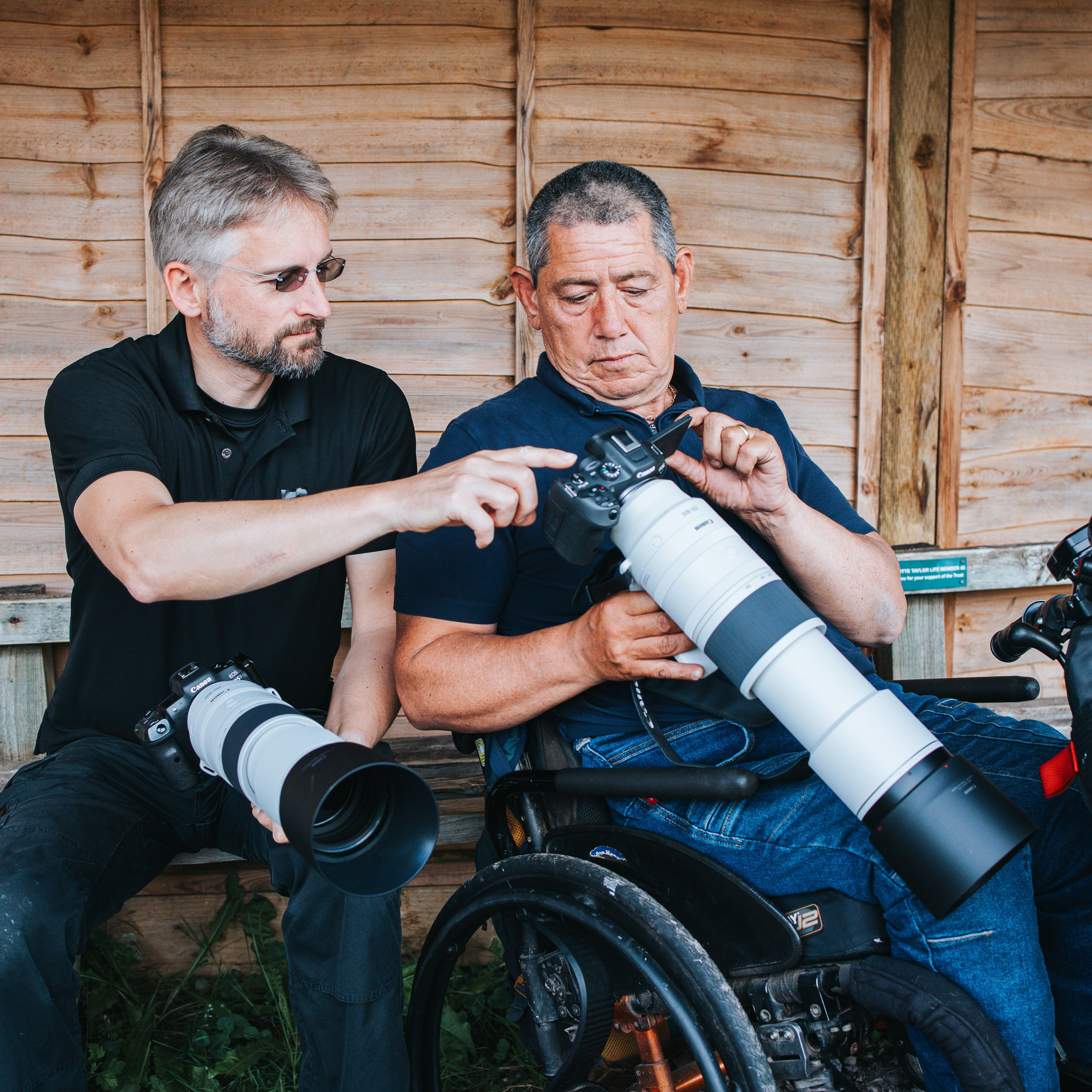 Deux hommes avec des appareils photo, un en fauteuil roulant. L'un pointe un appareil photo. Fond en bois. Longs objectifs.