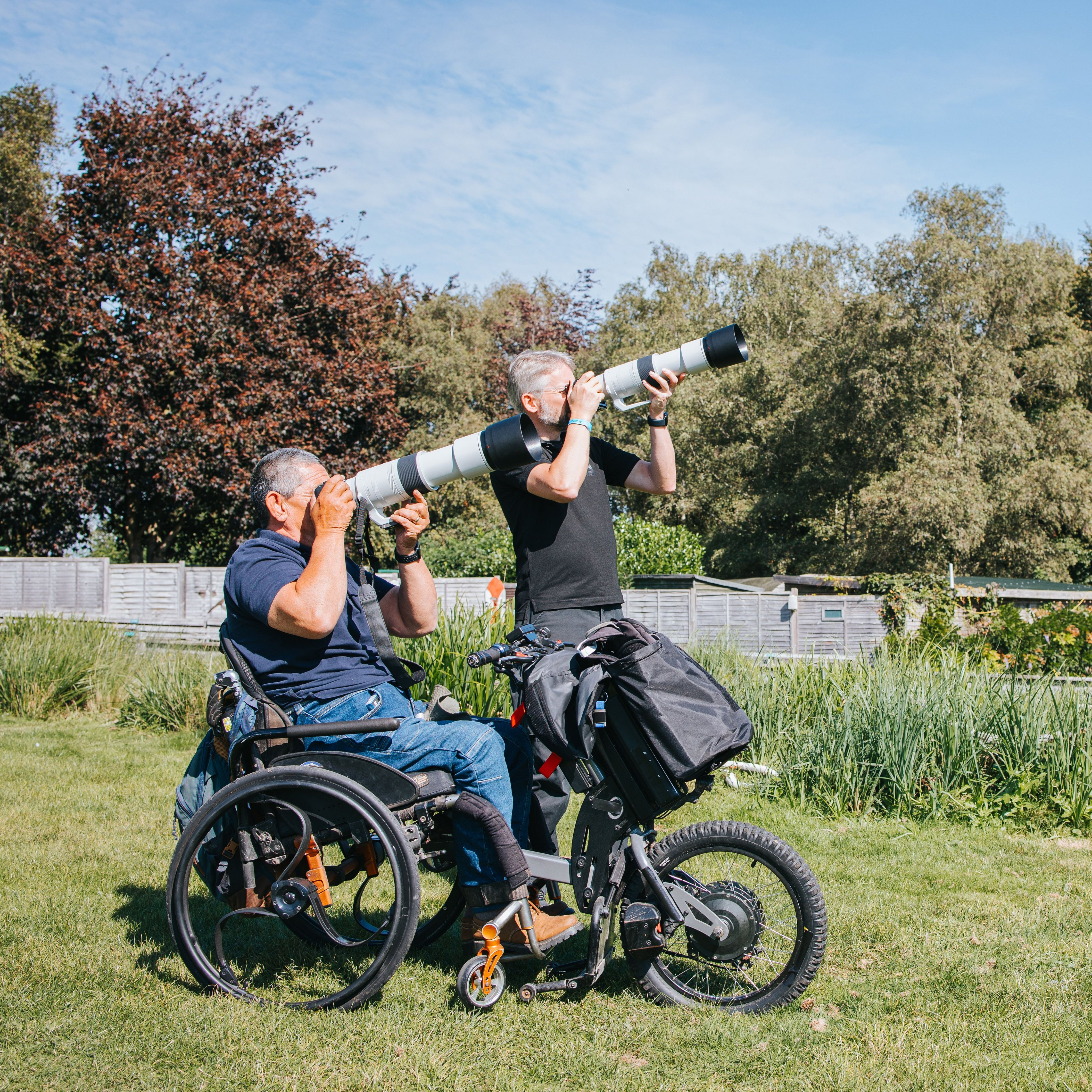 Deux hommes avec de grands objectifs en extérieur. Un en fauteuil roulant, l'autre debout. Herbe verte, arbres et ciel bleu en arrière-plan.