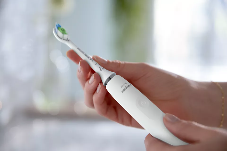 Mains tenant une brosse à dents électrique blanche. La brosse a des poils verts et bleus.
