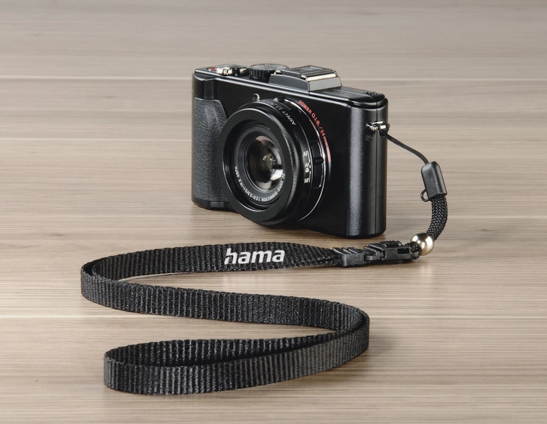 Appareil photo noir avec une sangle, sur une surface en bois, avec la marque 'hama'.