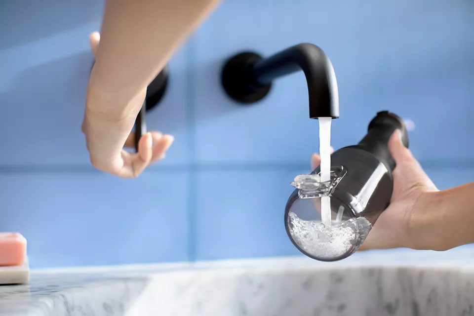 Mani che riempiono una bottiglia nera con acqua da un rubinetto nero.