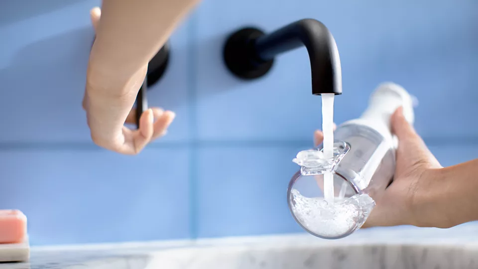 Mani di una persona che riempiono una bottiglia d'acqua trasparente da un rubinetto curvo.