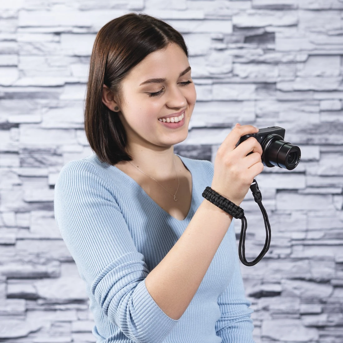 Une femme sourit en tenant un appareil photo noir. Elle porte une chemise bleue et une dragonne noire.