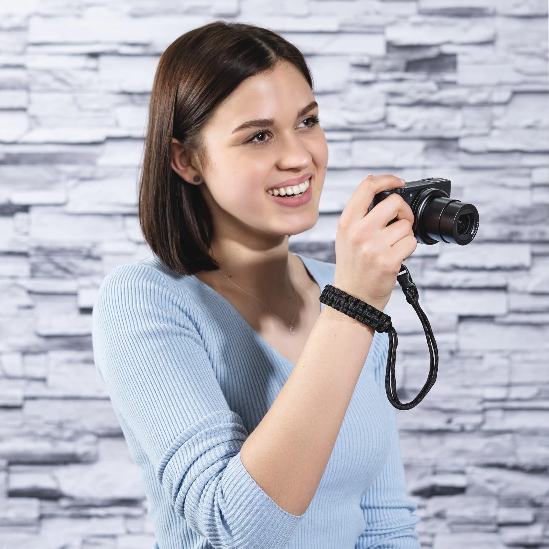 Femme souriante, tenant un appareil photo noir avec bracelet noir, contre mur de briques.