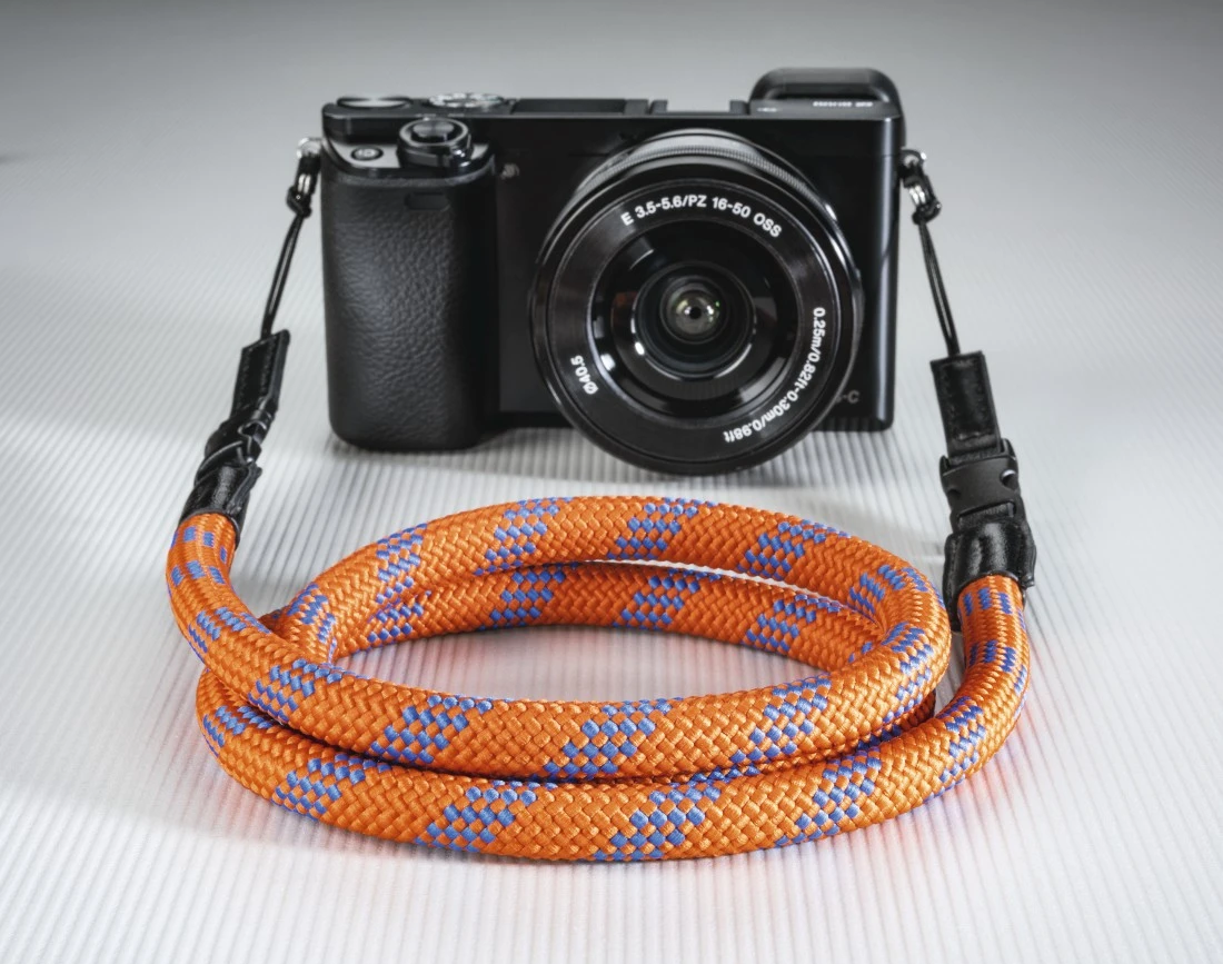 Una fotocamera con un cinturino arancione e blu è su una superficie bianca. Il cinturino è arrotolato e la fotocamera è nera.