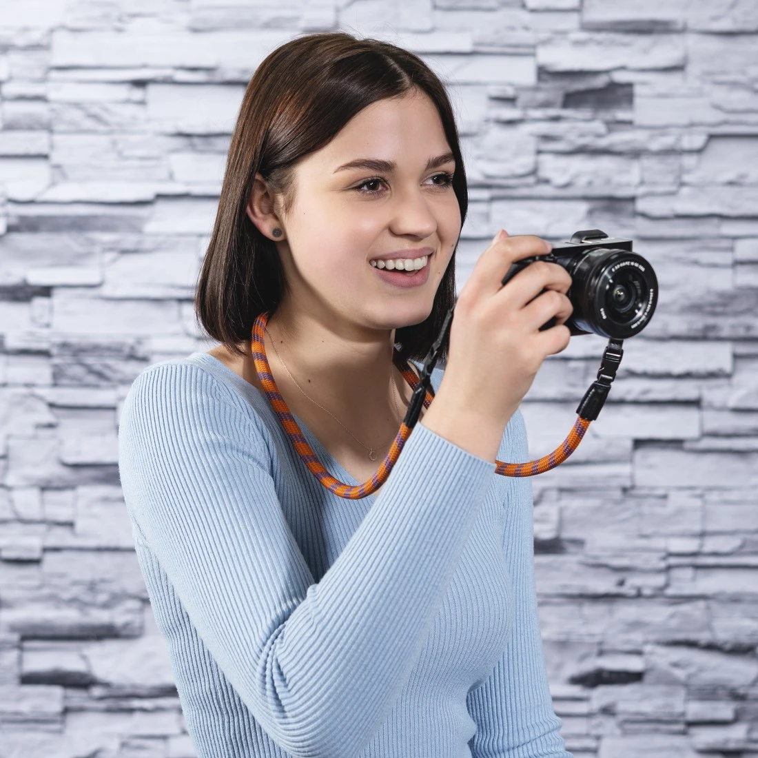 Donna sorride tenendo una fotocamera con una tracolla a righe arancioni e blu.
