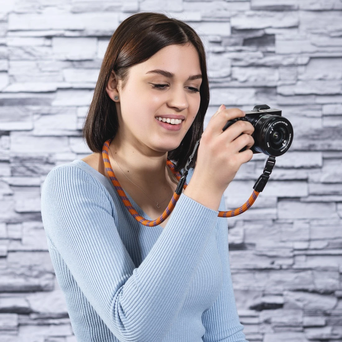 Una donna con una fotocamera al collo. Tiene la fotocamera, sorridendo. Il muro è grigio.