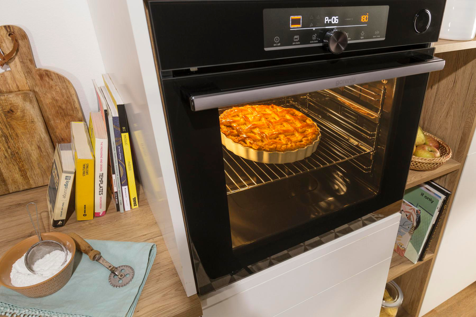 Ein Ofen mit einem Kuchen darin. Ein hölzernes Schneidebrett, Kochbücher und ein Mehl-Sieb sind an der Seite.