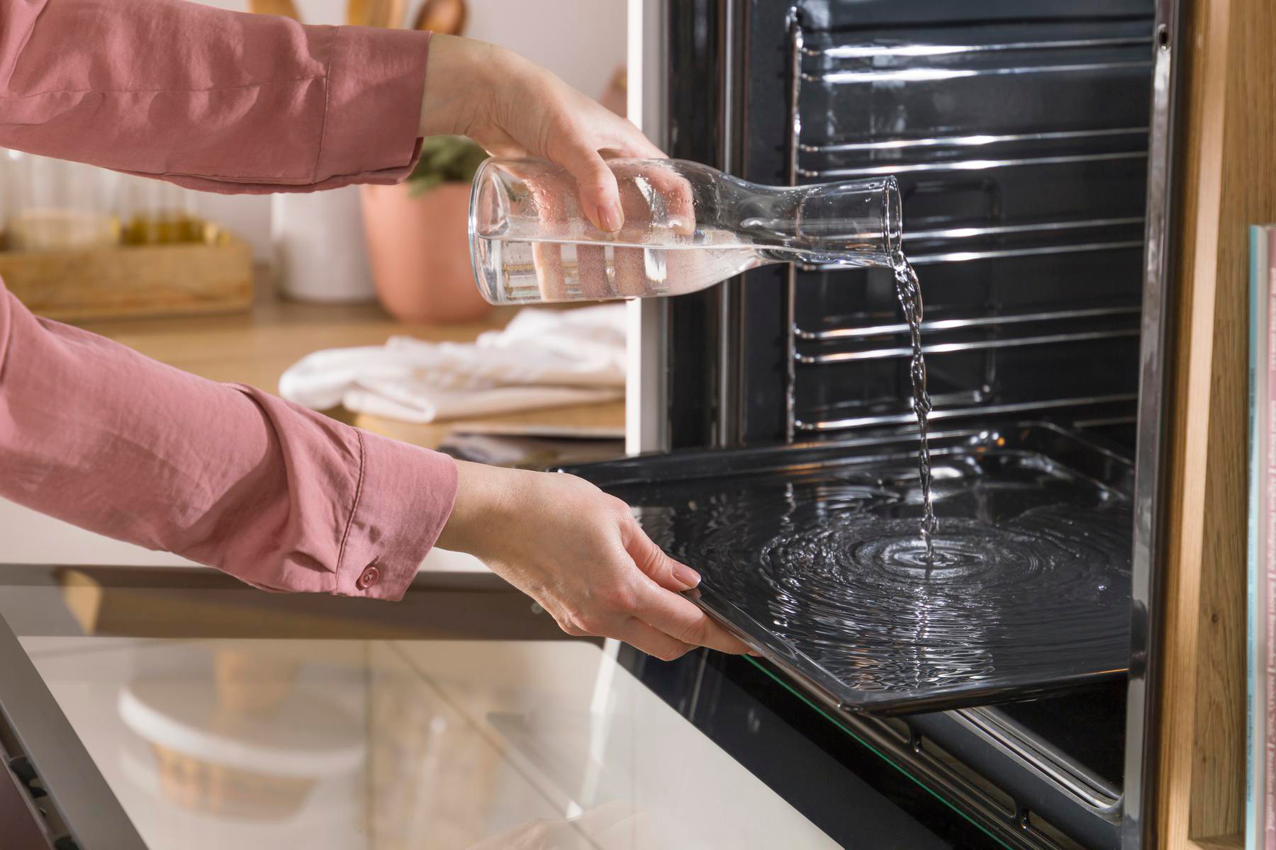 Frau gießt Wasser in ein Backblech in einem geöffneten Ofen. Küche im Hintergrund.