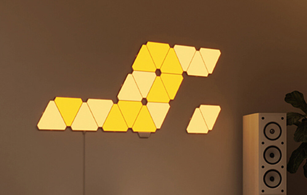 Geometryczna instalacja oświetlenia ściennego w żółtych tonach. Światło jest zamontowane na szarej ścianie.