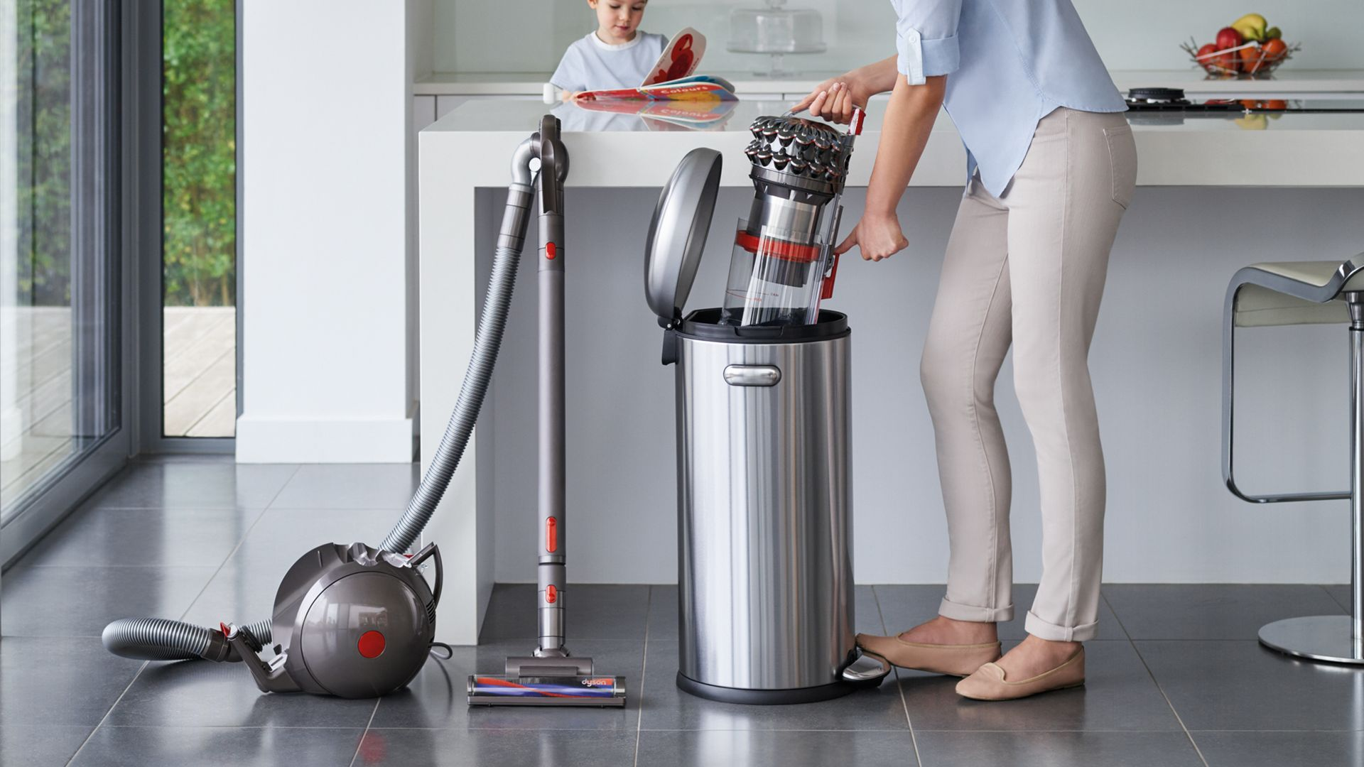 Bir kadın bir Dyson elektrikli süpürgeyi çöp kutusuna boşaltıyor. Arka planda bir çocuk görülüyor.