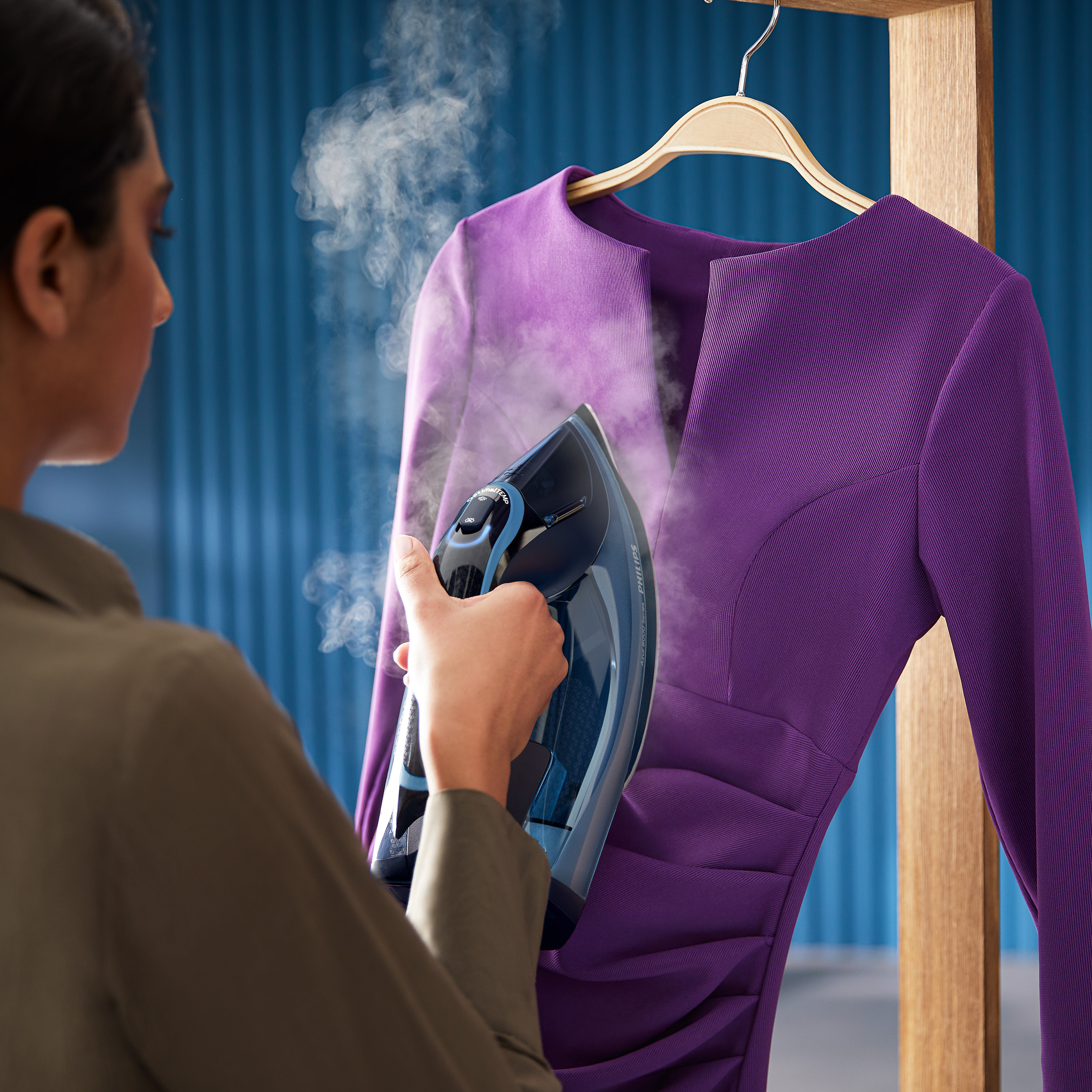 Frau, die ein violettes Kleid auf einem Holzgestell mit einem blauen Bügeleisen dämpft.
