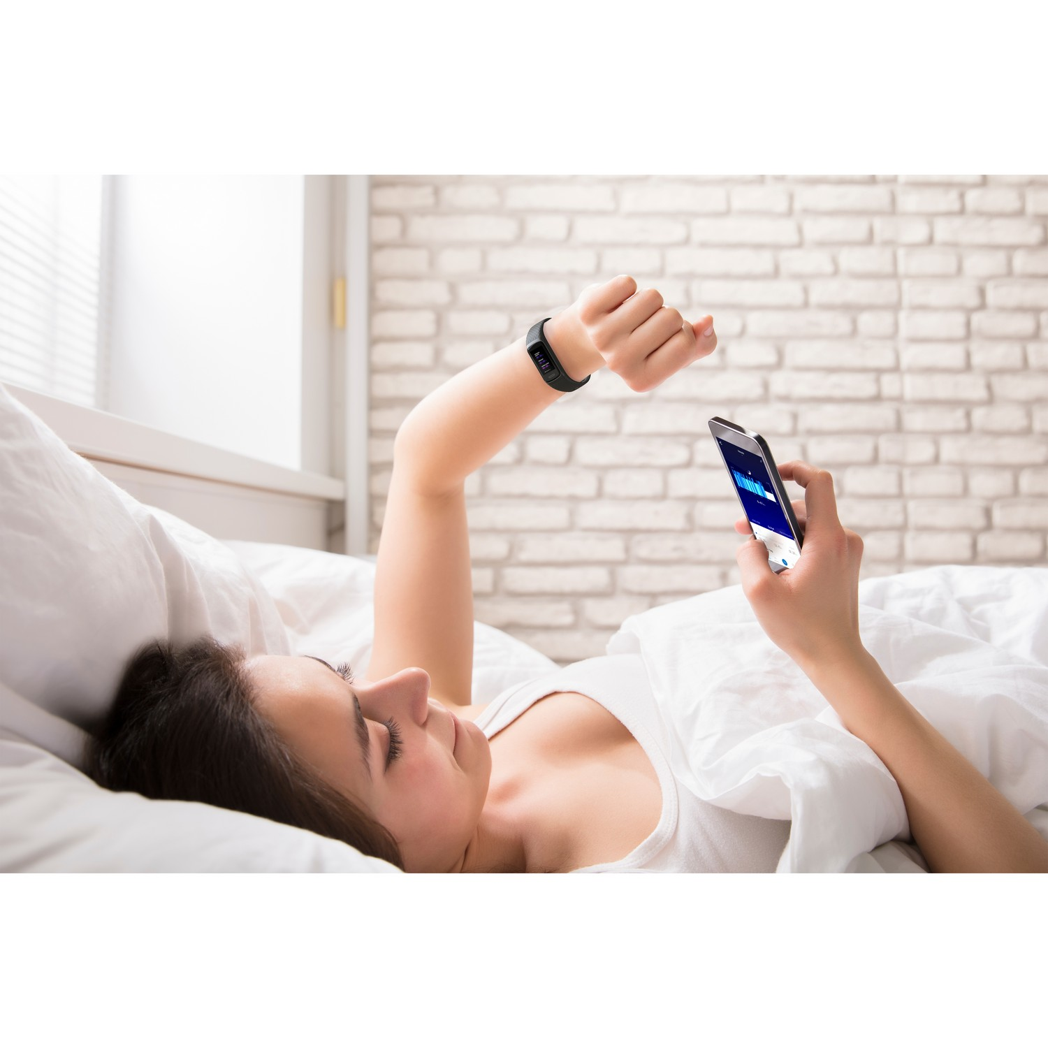 Une femme au lit vérifie son tracker de fitness à son poignet tout en tenant un téléphone.