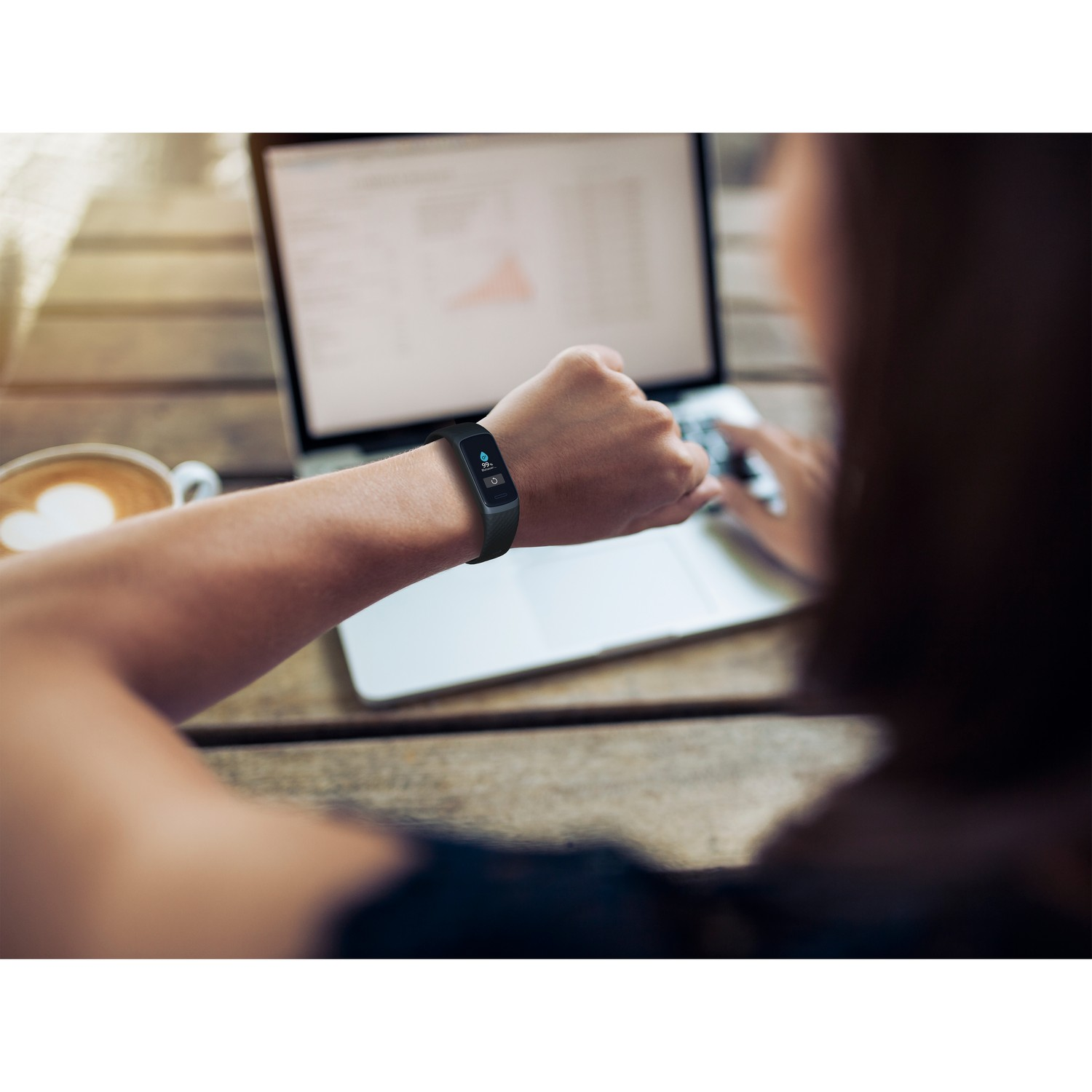 Une femme regarde une montre connectée à son poignet, assise à une table avec un ordinateur portable et du café.