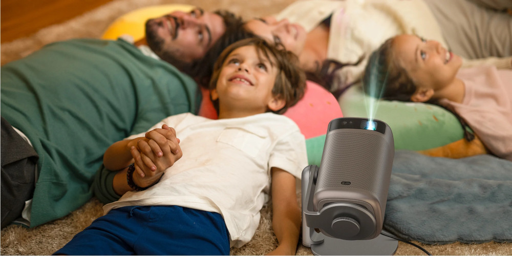 Een gezin ligt op een kleed en kijkt naar een projector die een film projecteert. Een projector staat op een standaard.