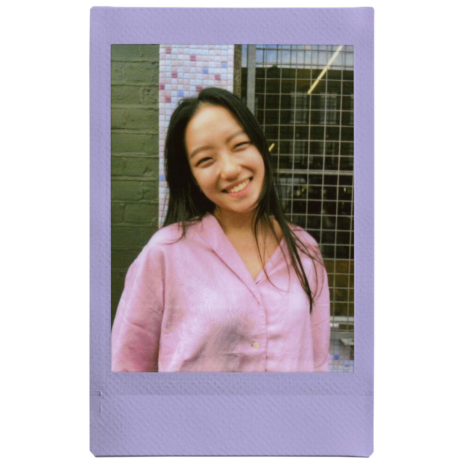 Photo polaroïd d'une femme asiatique souriante en chemise rose, devant un mur de briques et une clôture.