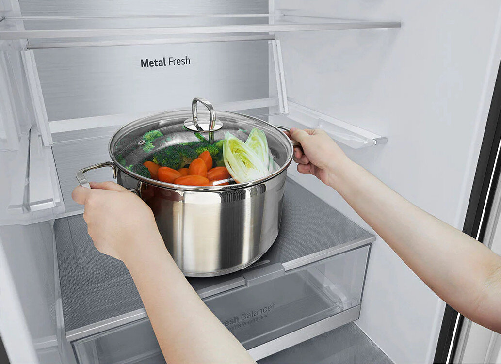 Ręce trzymające garnek z warzywami w lodówce. Metal Fresh to etykieta.